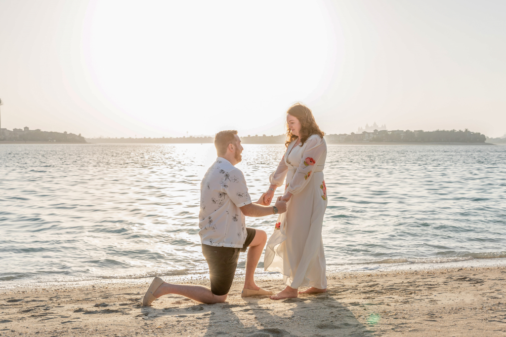Benji is down on one knee on the beach in Dubai holding both of Brogan's hands.